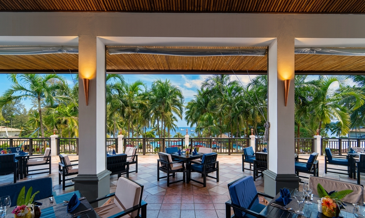 A view from a luxurious covered terrace dining area overlooking a pool, lush palm trees, and the blue ocean beyond.